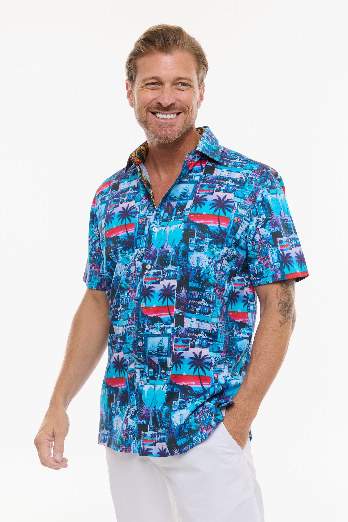 A smiling man wearing a short-sleeve button-up shirt with a colorful blue, red, and white tropical cityscape print, paired with white pants, against a plain white background.